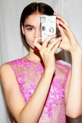 Model in a pink lace dress holding a vintage camera, posing at fashion week.