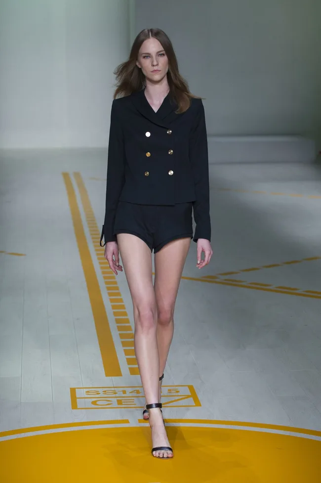 Model walking runway in black double-breasted jacket and shorts, with yellow lines on the floor.