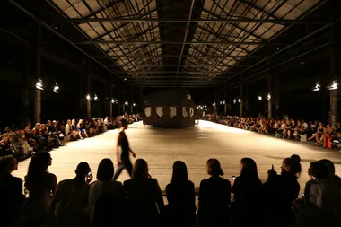 A runway show at Mercedes Benz Fashion Week with a large cabin structure center stage and audience seated on both sides.