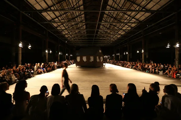 A runway show at Mercedes Benz Fashion Week with a large cabin structure center stage and audience seated on both sides.