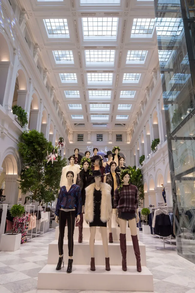 Mannequins in fashionable clothing displayed in an elegant, multi-story shopping mall with a glass ceiling.