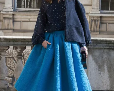 Woman in a polka dot blouse, blue textured midi skirt, and dark sunglasses standing against a brick building in daylight.