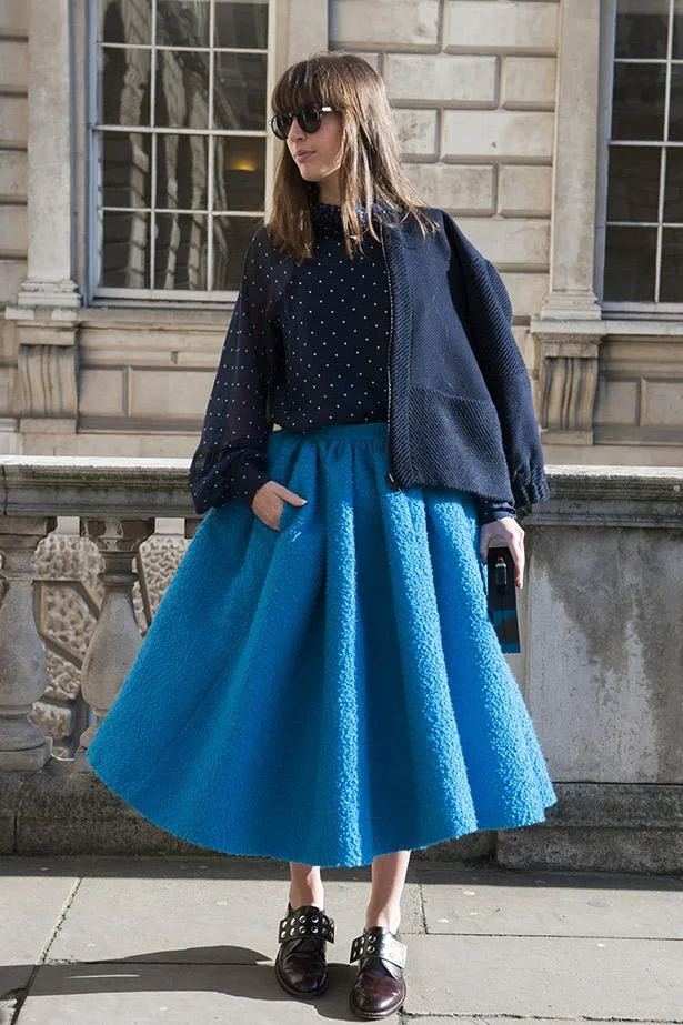 Woman in a polka dot blouse, blue textured midi skirt, and dark sunglasses standing against a brick building in daylight.