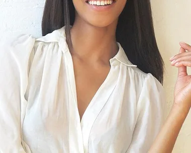 Woman with long straight hair in a white blouse smiling with hand raised against a white textured wall.