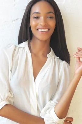 Woman with long straight hair in a white blouse smiling with hand raised against a white textured wall.