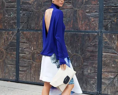 Woman in a blue backless top, holding a beige clutch, wearing white sneakers, with a patterned skirt and sunglasses.