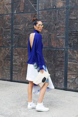 Woman in a blue backless top, holding a beige clutch, wearing white sneakers, with a patterned skirt and sunglasses.