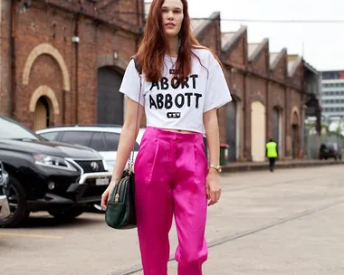 Woman in white "Abort Abbott" shirt, pink pants, black sneakers, with a green bag, standing on an urban street.