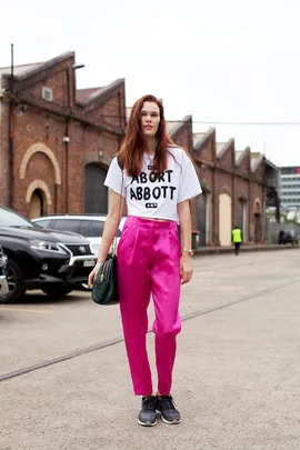 Woman in white "Abort Abbott" shirt, pink pants, black sneakers, with a green bag, standing on an urban street.