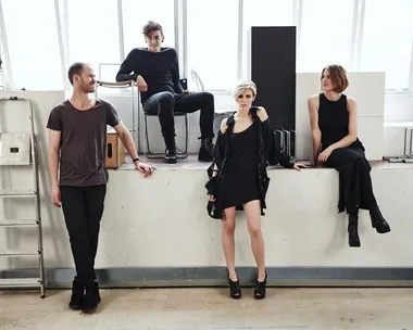 Four members of The Jezabels, a band, posing in casual clothing against a wall with bright windows behind them.