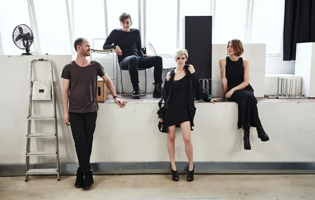 Four members of The Jezabels, a band, posing in casual clothing against a wall with bright windows behind them.