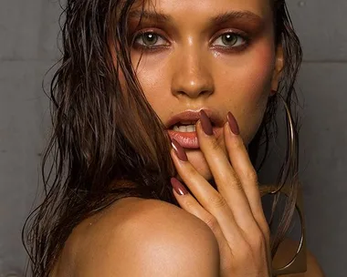 A woman with wet, slicked-back hair and brown eye makeup, holding her hand to her lips, showcasing long brown nails.