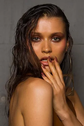 A woman with wet, slicked-back hair and brown eye makeup, holding her hand to her lips, showcasing long brown nails.