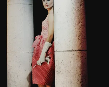 Audrey Hepburn in a pink dress and gloves, standing between large columns.