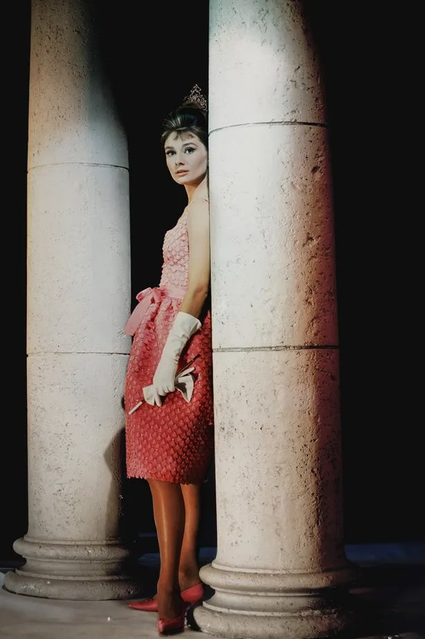 Audrey Hepburn in a pink dress and gloves, standing between large columns.