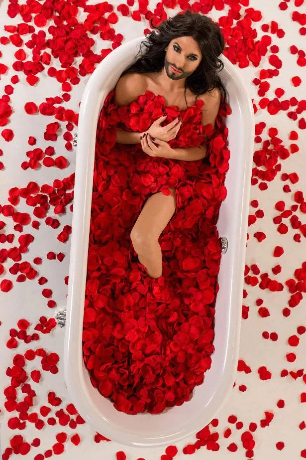 Person with long hair and beard in bathtub full of red rose petals, surrounded by more petals on floor.