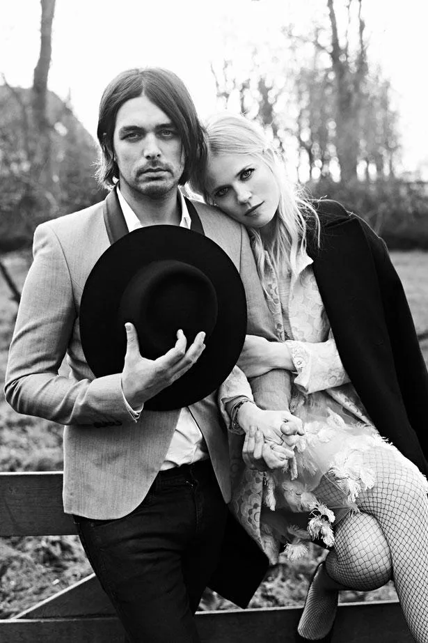 Man in a suit holding a hat and woman in a dress sitting on a fence, both in a black and white outdoor scene.