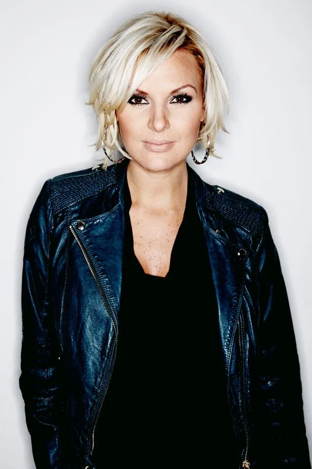 Blonde woman in a leather jacket, smiling slightly in a studio portrait.