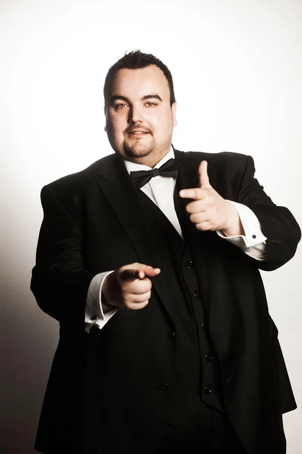 A man in a formal black tuxedo with a bow tie gestures with both hands, looking confident and smiling.
