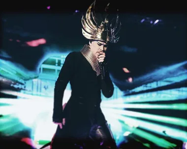 Performer in elaborate gold headdress sings on stage with vibrant lights at a music festival.