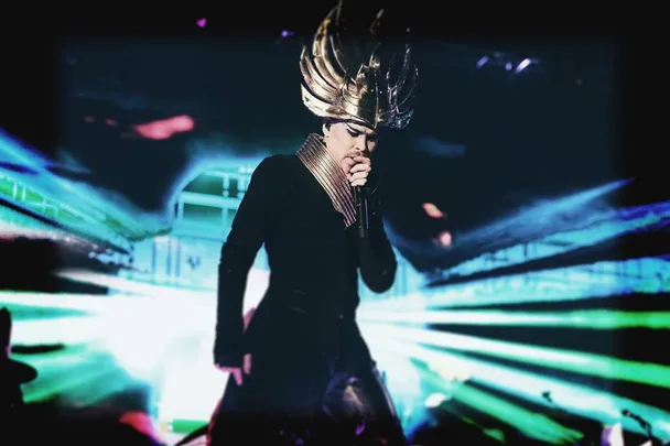 Performer in elaborate gold headdress sings on stage with vibrant lights at a music festival.