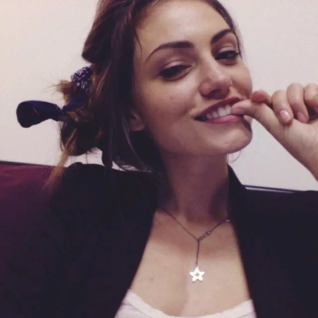 A woman smiling playfully with a flower necklace and a bun hairstyle, sitting indoors.