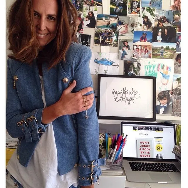 Woman in blue jacket smiling in front of photo-covered wall and desk with laptop displaying "Don't judge a book by its cover."
