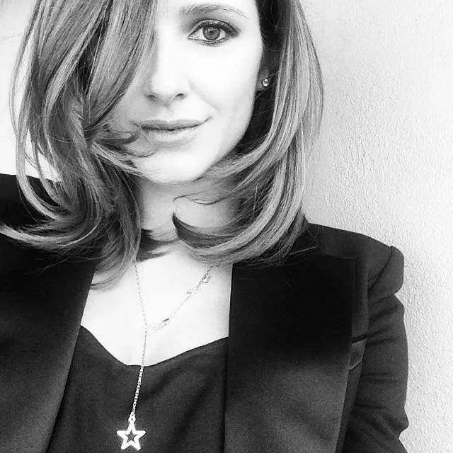 A woman with shoulder-length hair wearing a blazer and star necklace, captured in black and white, smiling slightly.