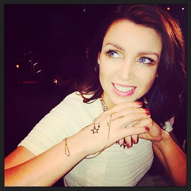 Celebrity woman smiling, wearing a star bracelet, with clasped hands against a dark background.