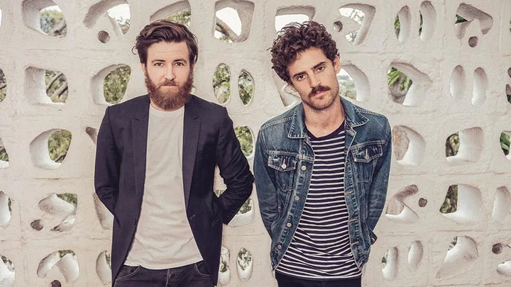 Two men pose in front of a white perforated wall; one in a blazer, the other in a denim jacket.