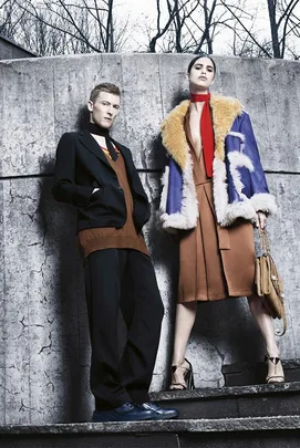 Models in stylish winter clothing pose dramatically against a concrete wall with a stark, leafless backdrop.