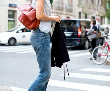 A woman in casual jeans, heels, and a scarf, crossing a city street with a red bag and a jacket.