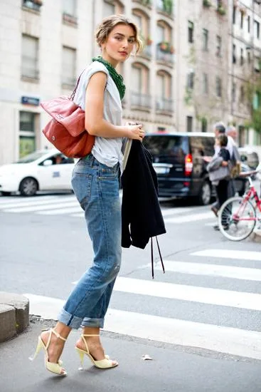 A woman in casual jeans, heels, and a scarf, crossing a city street with a red bag and a jacket.