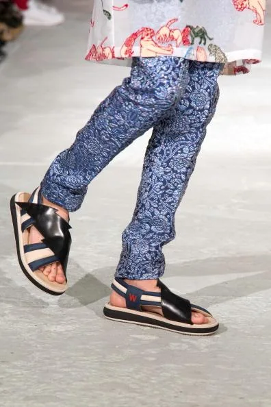 Person walking in floral-patterned trousers and black sandals on a runway.