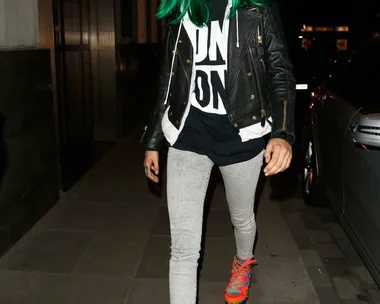 Person in green wig, black jacket, graphic tee, gray leggings, multicolored sneakers, and beanie walking on a city street.