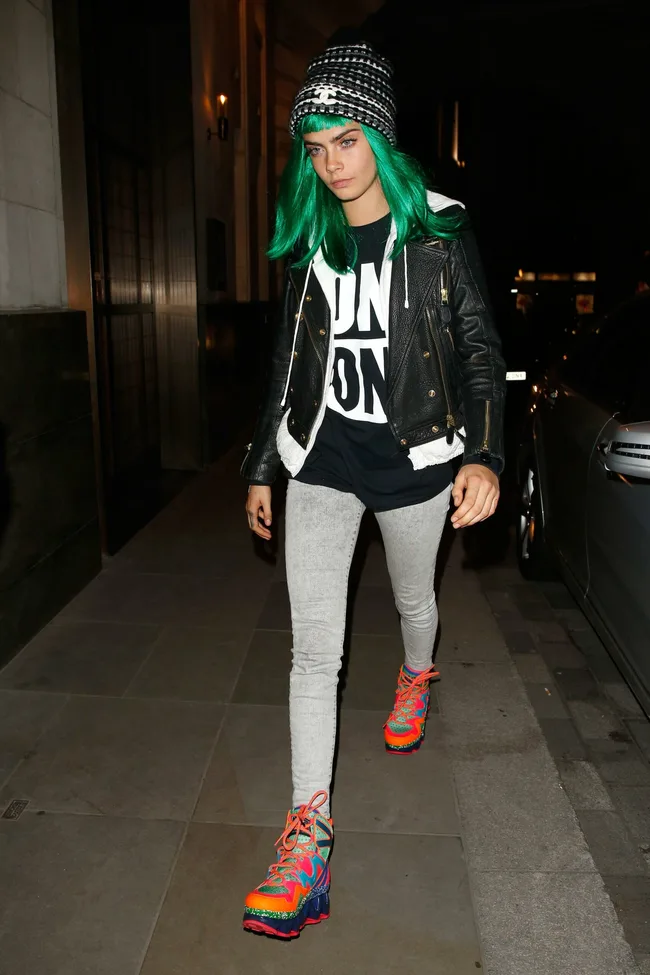 Person in green wig, black jacket, graphic tee, gray leggings, multicolored sneakers, and beanie walking on a city street.