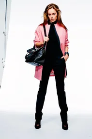 A woman in a pink coat, black outfit, and heels, holding a large black bag, posing confidently in a studio.