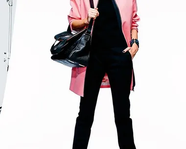 A woman in a pink coat, black outfit, and heels, holding a large black bag, posing confidently in a studio.