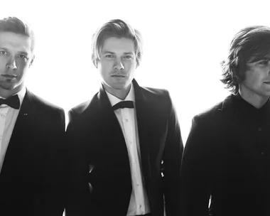 Black and white image of three men in suits and bow ties, standing side by side, with a bright backlight.