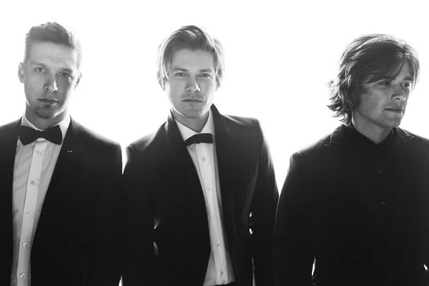 Black and white image of three men in suits and bow ties, standing side by side, with a bright backlight.