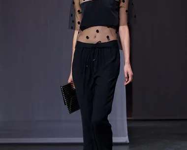 Model wearing sheer polka dot top and black pants on runway, holding a clutch.