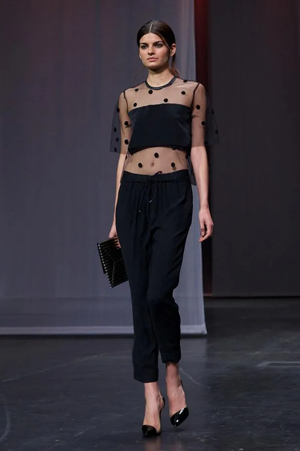 Model wearing sheer polka dot top and black pants on runway, holding a clutch.