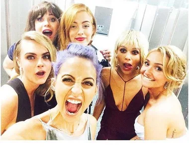 Group of six women taking a selfie, smiling and making playful expressions in a restroom setting.