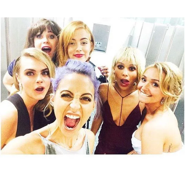 Group of six women taking a selfie, smiling and making playful expressions in a restroom setting.