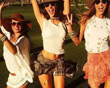 Three women in festival attire with hats and sunglasses, posing happily on a sunny day with palm trees in the background.