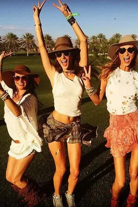 Three women in festival attire with hats and sunglasses, posing happily on a sunny day with palm trees in the background.