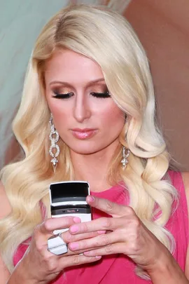 Blonde woman with long hair using a flip phone, wearing a pink outfit and chandelier earrings.