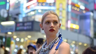 A woman with blonde hair and red lipstick, adorned with a pearl necklace, stands in a brightly lit urban setting, possibly Tokyo.