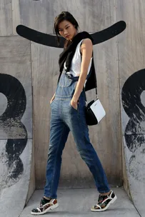 A person wearing denim overalls, a white top, and a black sweater, poses in front of a wooden wall with a black graffiti design.