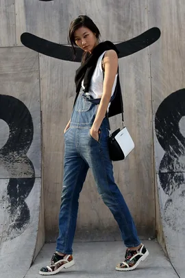 A person wearing denim overalls, a white top, and a black sweater, poses in front of a wooden wall with a black graffiti design.